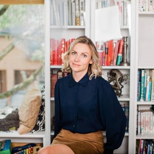 Eva Jurczyk, hands in pockets, sits on a stool in front of bookshelves.