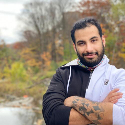 Faraz Khoshbakhtian smiles, standing by a river in a wooded area.