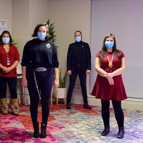 Five people wearing masks, and standing 2 metres apart, pose for a picture on a cosy, colourful carpet.