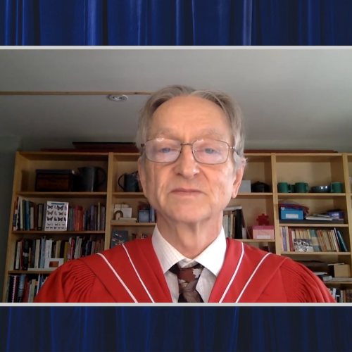 Geoffrey Hinton, wearing academic robes, stands in front of a bookcase and speaks on video.