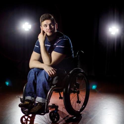 George Alevizos, looking thoughtful, sits on a stage with spotlights behind him.
