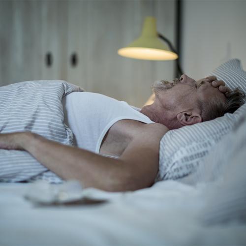 A man lies on his back in bed with his eyes closed and one hand on his head.