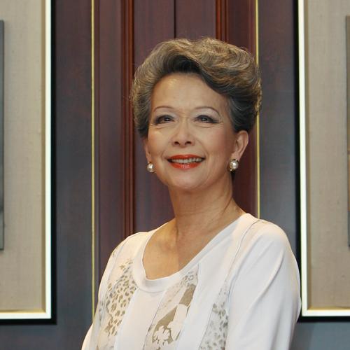 Vivienne Poy smiles, sittingin a room with artwork on the walls showing people in traditional Chinese dress.