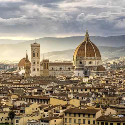 A medieval cathedral decorated in a tile pattern soars above the tiled rooftops of Florence at sunset.