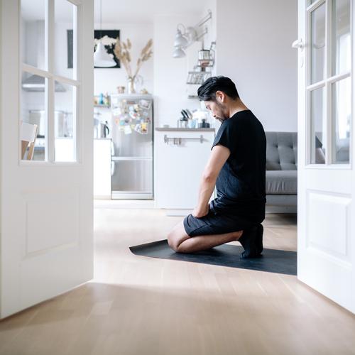 A man kneels upright, head slightly bowed and eyes closed, on the floor of a sunny room.