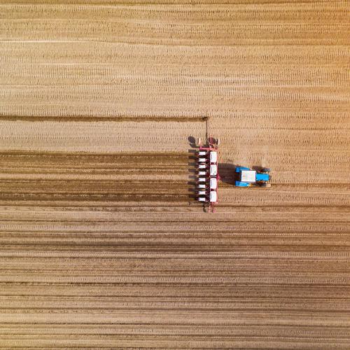 Seen from high above, a tractor drags a machine across a crop field, leaving stripes.