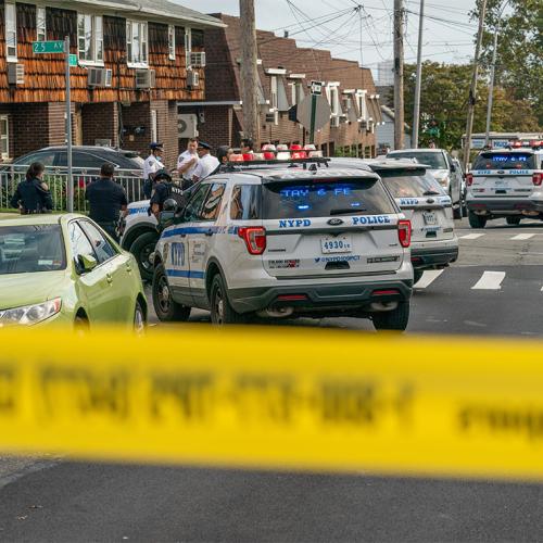 Police tape blocks off a residential street. Behind the line, police gather around their parked cars and talk seriously.