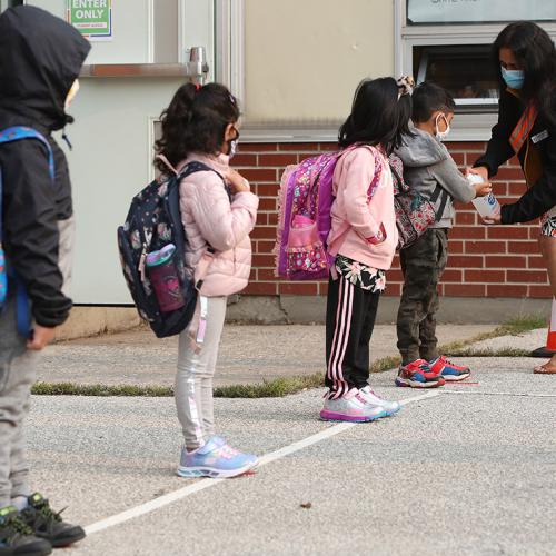 Four small children, wearing masks, stand spaced apart in a playground while a teacher in a mask gives them hand sanitizer.