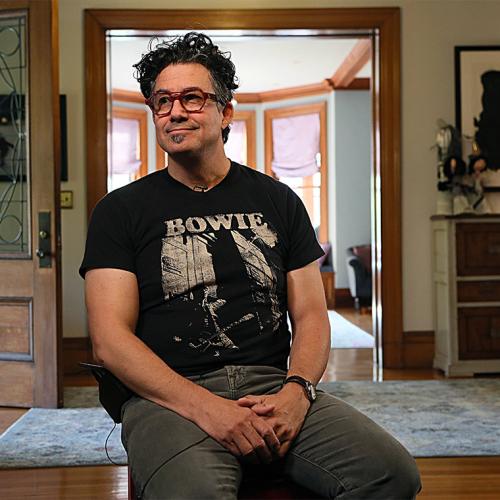 Derek Rossi, wearing a Bowie T-shirt and headphones, smiles as he sits in the hallway of a large house.