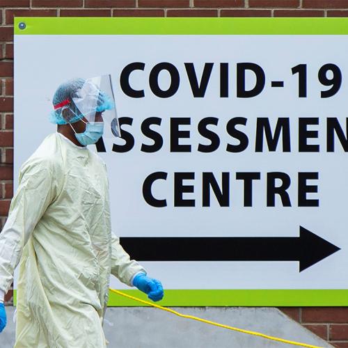 A person in full protective equipment walks by a sign that reads COVID-19 Assessment Centre.