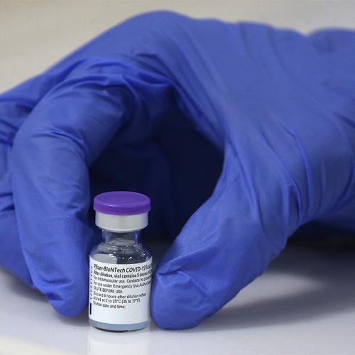 A hand in a medical glove holds a tiny vial. The vial is labelled: Pfizer-BioNTech COVID-19 vaccine.