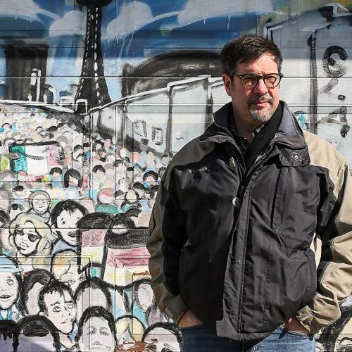 Colin Furness stands in front of a wall mural that shows a crowded streetcar platform.