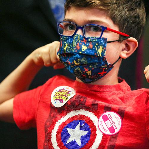 A young boy flexes his muscles. He's wearing a mask, a superhero t-shirt, and a sticker that says I got my COVID-19 vaccine.