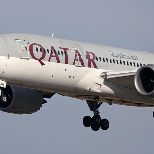 A jumbo jet with wheels down flies low. Letters on the side of the plane read: Qatar.