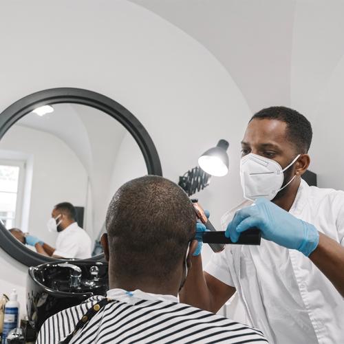 A Black barber in mask and gloves puts the finishing touches on a haircut for a man sitting in a barber chair.