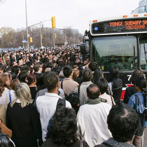 In a pre-pandemic photograph, hundreds of people crowd towards the doors of one lone bus labelled Yonge Subway Shuttle..