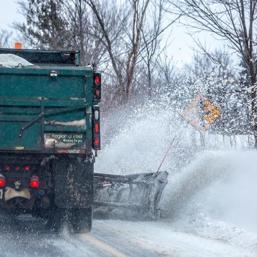 A salt truck with plow sends snow flying over a snowbank at the side of a tree-lined rural road.