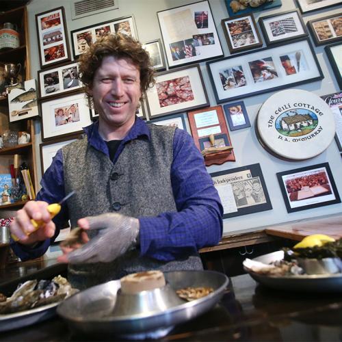 Patrick McMurray smiles as he shucks an oyster at a wooden table near a wall covered with photos and newspaper clippings.