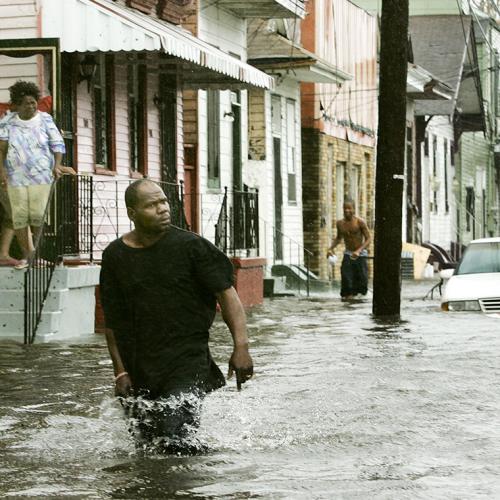 A man wades through knee-deep water down a residential street while people look out their doors.