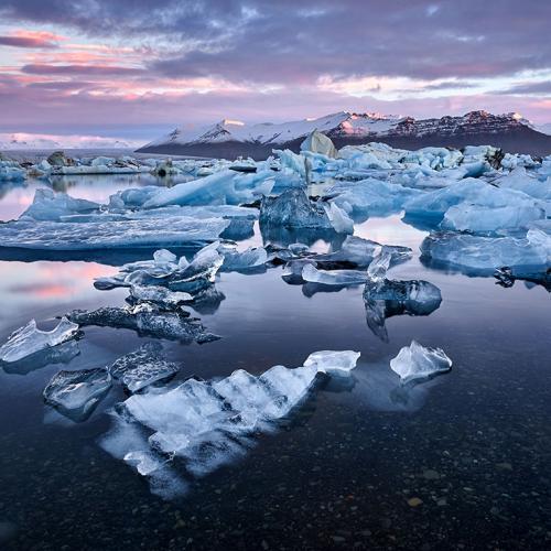 Melting ice floats in a glass-calm Arctic inlet, with snow-covered mountains in the background.