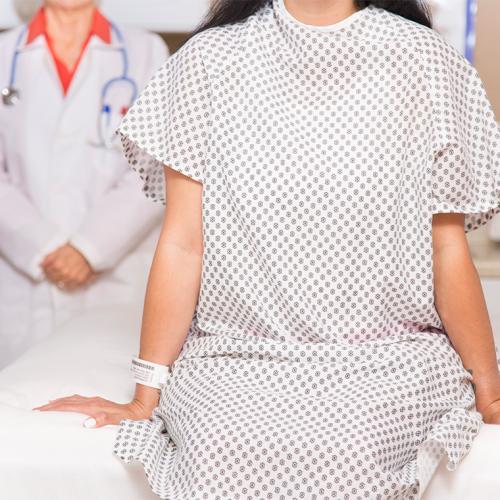 A white woman sits on a hospital bed in a hospital gown, while a figure in a white coat stands behind.