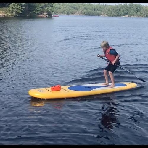 On a lake, a boy stands and jumps on the stern of a paddleboard. Ripples behind show he's moving forward.