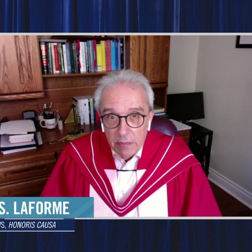 Harry LaForme, wearing his academic robes, speaks on video in front of a wooden desk and bookshelf.
