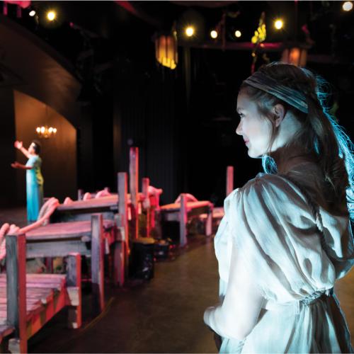 A woman in costume watches from the back of the Hart House Theatre stage as a woman in the spotlight holds up her arms.