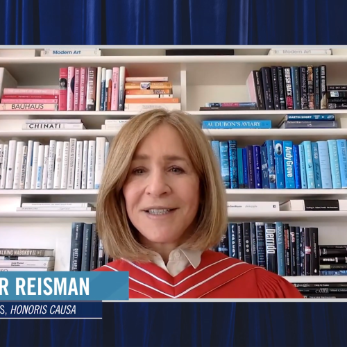 Heather Reisman, wearing academic robes, speaks on video in front of a bookcase of books arranged by colour.