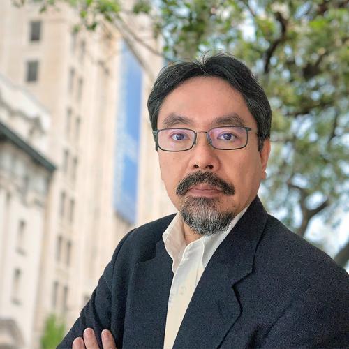 Naoto Hirano looks serious as he stands under a tree outdoors.