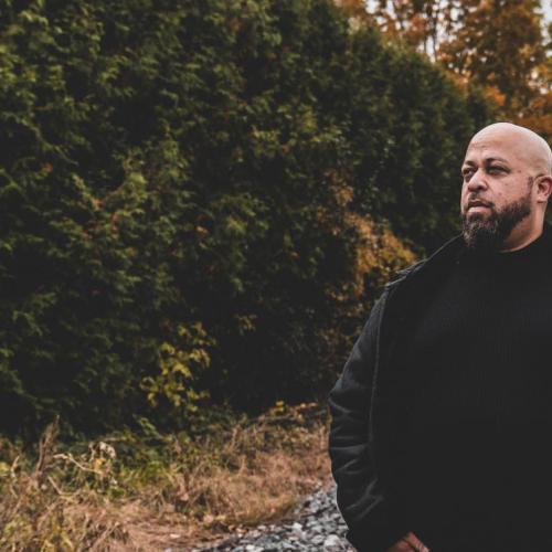 Ian Andre Espinet looks thoughtful, standing by a hedge outdoors.