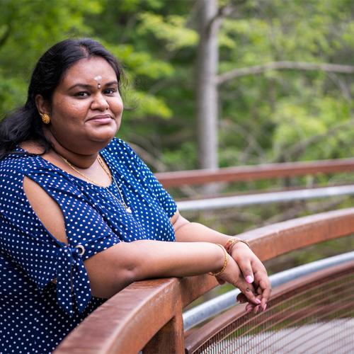Ilakkiah Chandran smiles, leaning on a railing on a boardwalk in the woods.