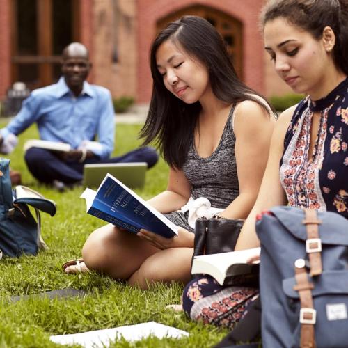 Two female alumni sitting together reading in the grass