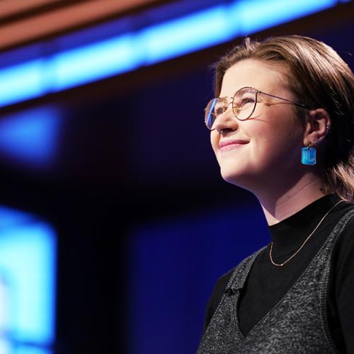 Mattea Roach smiles happily, standing in the lights on the Jeopardy TV set.
