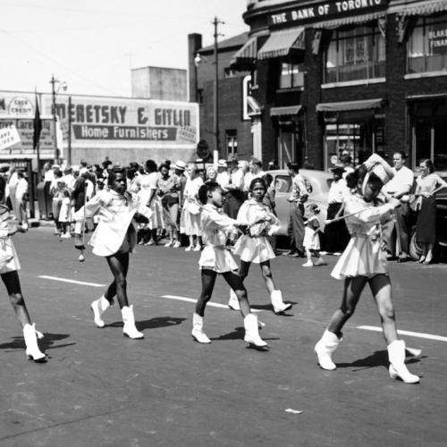 In an undated old photograph, young Black girls in majorette costumes march along a street lined with smiling white people.