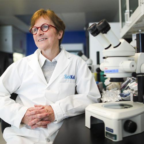 Janet Rossant smiles, wearing a Sick Kids white medical coat and sitting next to a microscope on a bench.