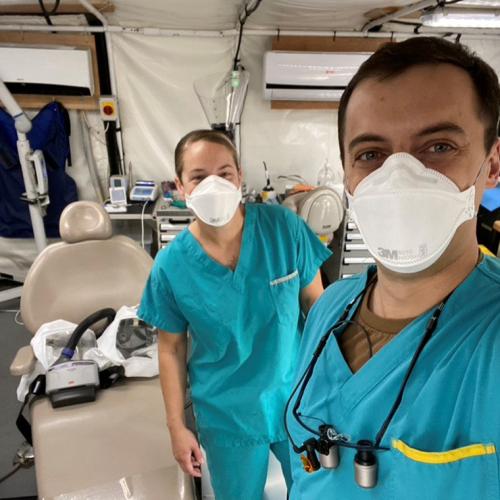 Sgt. Angela Brownell and Jesse Barker, wearing scrubs and masks, stand beside a dental chair inside a large tent.