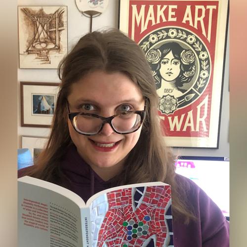 Jessica Watkin holds up an open book. Behind her, a poster that says, make art not war. 