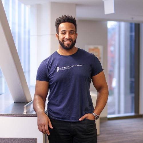 Joshua Lokko smiles, wearing a U of T Faculty of Law T-shirt and standing by an indoor balcony railing.