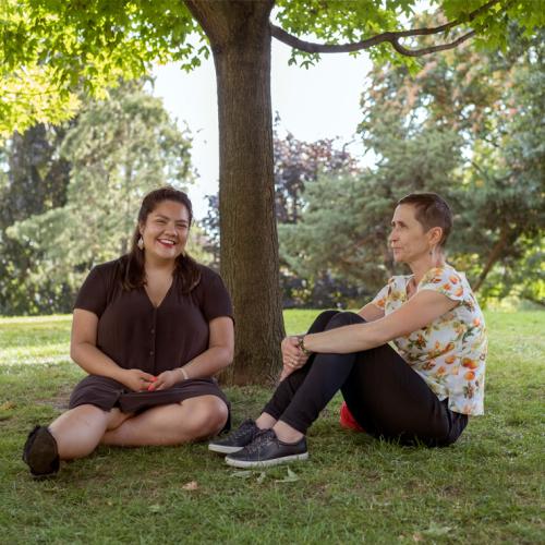 Kateri Lucier-Laboucan and Andrea Mantin smile together, sitting on a lawn under a tree.