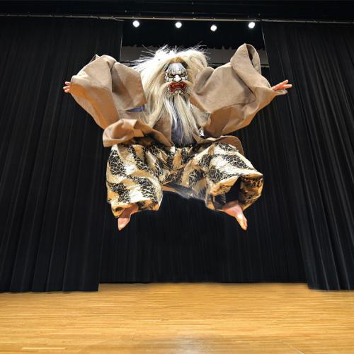 A masked performer jumps up on stage with black drapes behind him