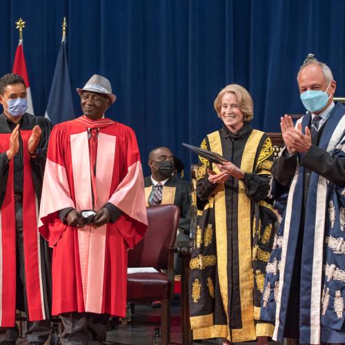 Meric Gertler and Rose Patten applaud Winston LaRose on stage in Convocation Hall. All three wear academic robes.