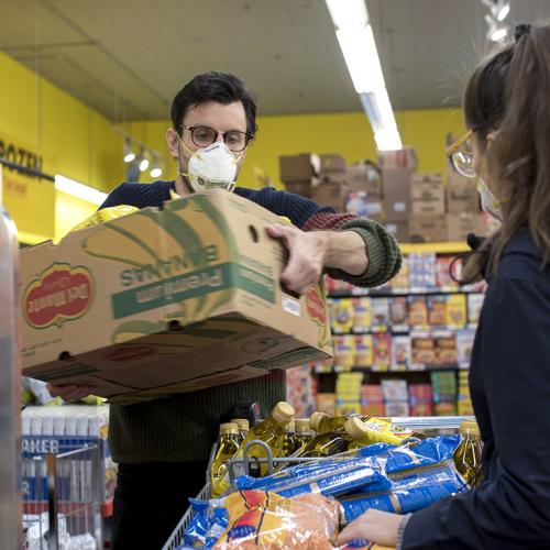 In a grocery store, Adam Zivo lifts a cardboard box full of groceries. He is wearing a mask.