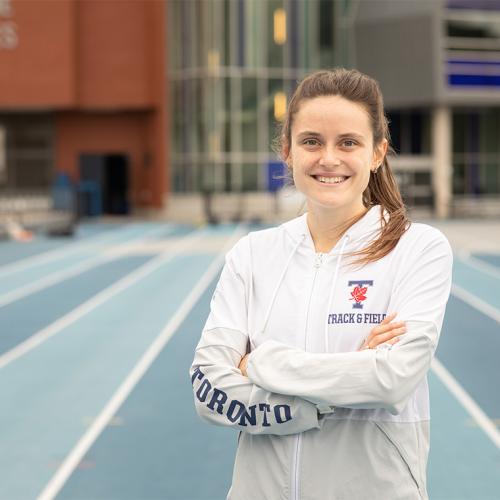 Lucia Stafford smiles as she stands with arms crossed on the Varsity Stadium track. She wears a Varsity Blues hoodie.