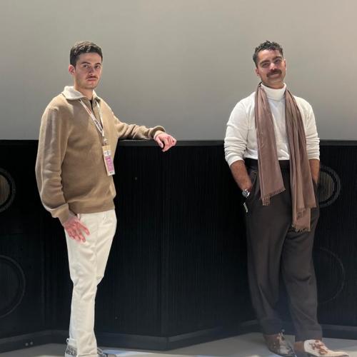 A portrait of two people standing next to waist-high black speakers