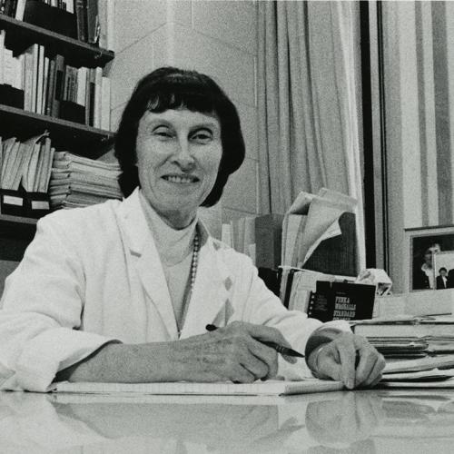 Marian Packham smiles as she sits at an office desk, wearing a lab coat.