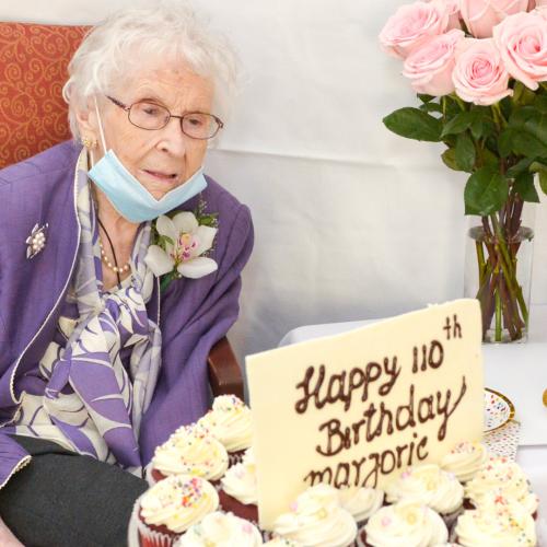 Marjorie Douglas, wearing a flower in her buttonhole, smiles at a tray of cupcakes and a sign saying Happy 110th Birthday.