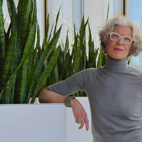 A woman in a grey turtleneck and glasses poses next to some potted plants