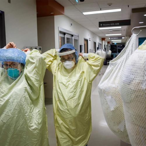 Two masked medical students lift their arms and tie their protective gowns behind their necks as they walk down a hallway.
