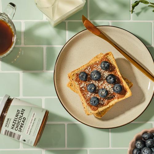 A slice of toast with chocolate spread and blueberries, beside a jar labelled Mounib Hazelnut Chocolate Spread.
